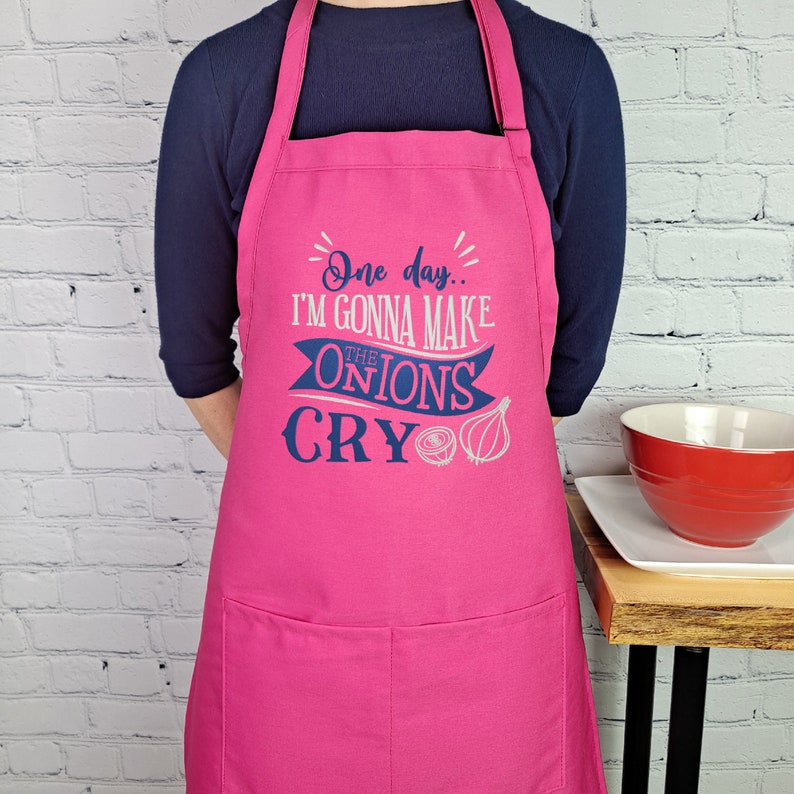 May include: Pink kitchen apron with a blue and white design that says "One day... I'm gonna make the onions cry".