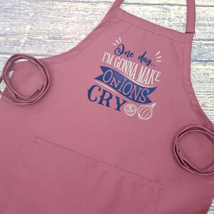 May include: A maroon kitchen apron with a pocket and adjustable straps. The apron features a white and blue graphic that says "One day... I'm gonna make the onions cry".
