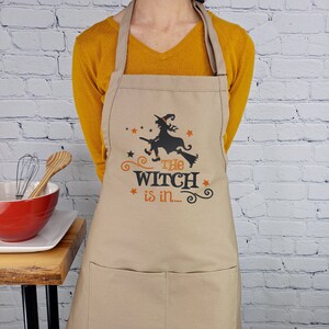 Puede incluir: Un delantal de cocina beige con una silueta negra de una bruja volando en una escoba. El texto "The Witch is in..." est&aacute; impreso en naranja en el delantal.