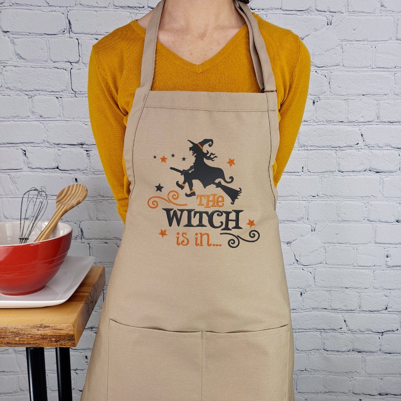 Puede incluir: Un delantal de cocina beige con una silueta negra de una bruja volando en una escoba. El texto "The Witch is in..." est&aacute; impreso en naranja en el delantal.