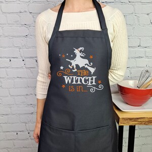 Puede incluir: Delantal de cocina negro con una silueta blanca de una bruja volando en una escoba. El texto "The Witch is in..." est&aacute; impreso en naranja debajo de la bruja.