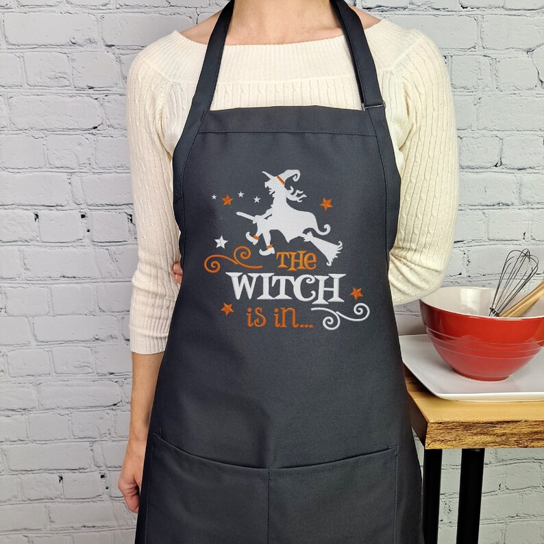 Puede incluir: Delantal de cocina negro con una silueta blanca de una bruja volando en una escoba. El texto "The Witch is in..." est&aacute; impreso en naranja debajo de la bruja.