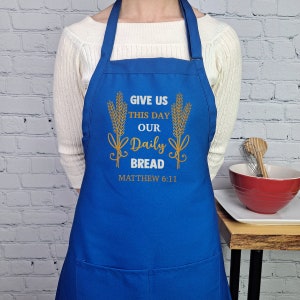 Op de afbeelding: Blauwe keukenschort met een gouden ontwerp met de tekst "Geef ons heden ons dagelijks brood" en "Mattheüs 6:11".