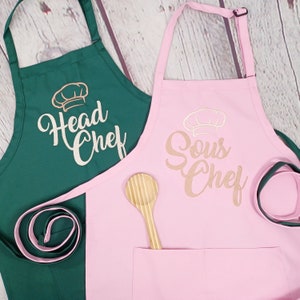 May include: Two aprons, one green and one pink, with the text "Head Chef" and "Sous Chef" embroidered on them. Both aprons feature a chef's hat design.