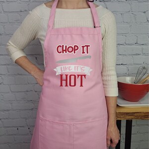 Pu&ograve; includere: Grembiule da cucina rosa con un'immagine di coltello bianco e il testo "Chop it like it's hot" in rosso.