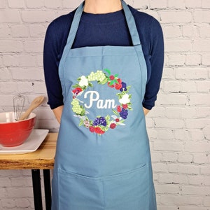 May include: A blue denim apron with a pocket and adjustable straps. The apron has an embroidered wreath of berries and flowers with the name "Pam" in the center.