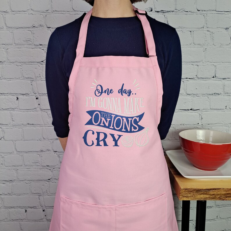 May include: Pink kitchen apron with a blue and white design that says "One day... I'm gonna make the onions cry".