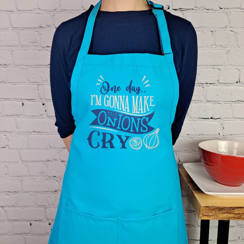 May include: A turquoise blue apron with adjustable straps and a large pocket. The apron features a white and blue design that says "One day... I'm gonna make the onions cry".