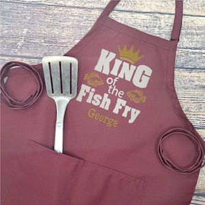 May include: Burgundy apron with a white embroidered design that says "King of the Fish Fry George". The design also includes a gold crown and two fish.