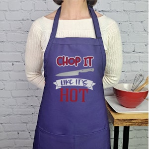 Pu&ograve; includere: Grembiule da cucina viola con una grafica bianca e rossa che dice "Chop it like it's hot" con una grafica di coltello sopra il testo.