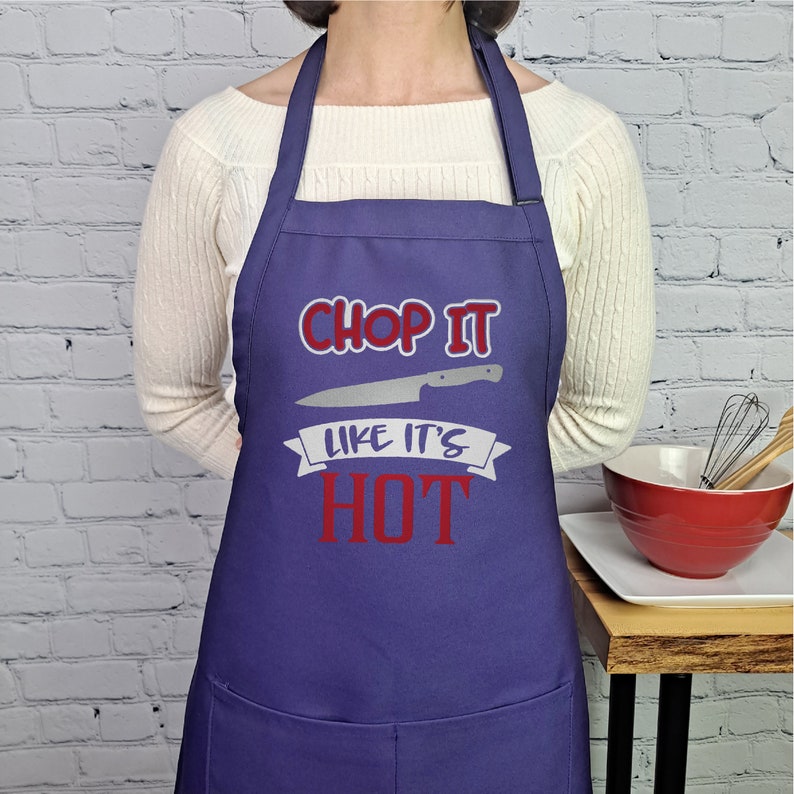 Pu&ograve; includere: Grembiule da cucina viola con una grafica bianca e rossa che dice "Chop it like it's hot" con una grafica di coltello sopra il testo.