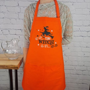 Puede incluir: Delantal de cocina naranja con una silueta negra de una bruja volando en una escoba. El texto "The Witch is in..." est&aacute; impreso en el delantal.
