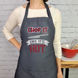 Pu&ograve; includere: Un grembiule grigio con un design rosso e bianco che dice "Chop it like it's hot". Il grembiule ha una tasca sul fondo e un grafico di un coltello.