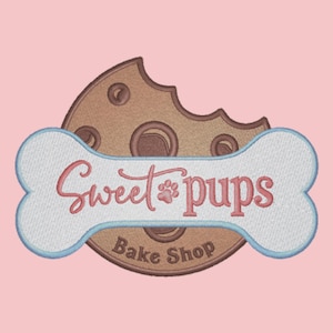 May include: A brown cookie with a bite taken out of it, with a white bone-shaped banner that says "Sweet Pups Bake Shop" in pink and red lettering. The cookie has a paw print in the center of the banner.