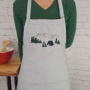 May include: A light blue apron with a pocket and an embroidered design of a mountain range, a tent, a campfire, and trees.