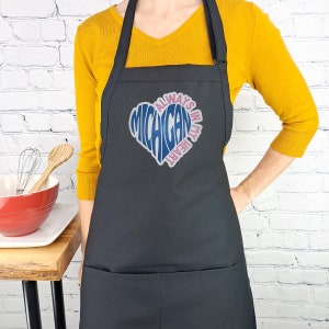 Op de afbeelding: Zwarte schort met een roze en blauw hartontwerp met de tekst "Always in my heart Michigan".