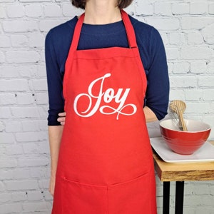 May include: Red kitchen apron with a white embroidered word "Joy" on the front. The apron has a large pocket on the bottom.