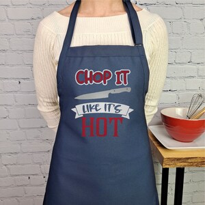 Pu&ograve; includere: Un grembiule blu navy con il testo "Chop it like it's hot" in rosso e bianco. Un coltello argentato &egrave; raffigurato sopra il testo.