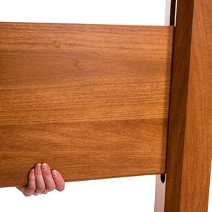 May include: Close-up of a wooden bed frame component. The wood has a warm, reddish-brown stain and visible grain. A hand is holding the frame, suggesting assembly or inspection. The design appears to be a modern style.