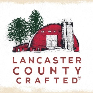 May include: A red barn with a white silo, surrounded by snow and two green trees. The text "LANCASTER COUNTY CRAFTED" is below the barn in red.