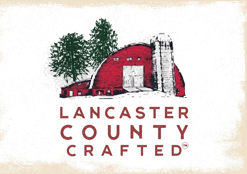 May include: A red barn with a white silo, surrounded by snow and two green trees. The text "LANCASTER COUNTY CRAFTED" is below the barn in red.