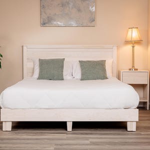 May include: A white wooden bed frame with a horizontal plank headboard, featuring a white quilted comforter and two sage green square throw pillows. The bed is positioned in a room with a light-colored wall, a bedside table, and a lamp.