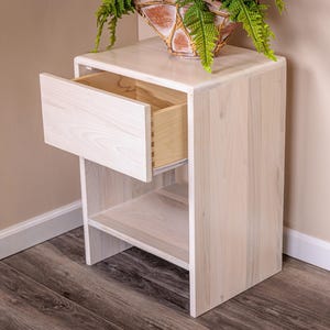 May include: A light-colored wooden nightstand featuring a single pull-out drawer with visible dovetail joinery on the side and an open storage shelf underneath. The wood grain is light and natural, and the drawer is partially extended outward.