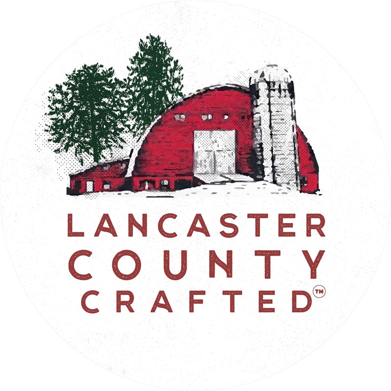 May include: A red barn with a white door and a silo in a snowy landscape. The text "Lancaster County Crafted" is below the barn in red letters with a trademark symbol.