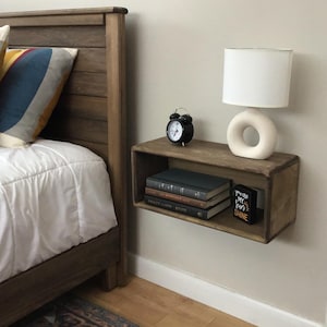 May include: A wooden floating shelf with a black alarm clock, a white lamp with a ring base, and books. The shelf is mounted on a white wall next to a bed with a wooden headboard.