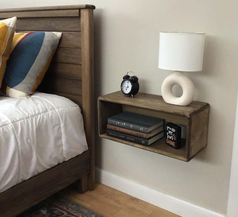 May include: A wooden floating shelf with a black alarm clock, a white lamp with a ring base, and books. The shelf is mounted on a white wall next to a bed with a wooden headboard.