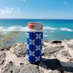May include: A blue and white patterned can cooler holds a can of juice. The cooler has a plaid design with white floral accents. The background features a rocky surface, the ocean, and a bright blue sky with clouds.