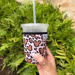 以下が含まれることがあります： 茶色のミッキーマウス柄のピンク色のドリンクスリーブ。ストローの付いた透明なプラスチックカップを包んでいます。スリーブの上部には黒いバンドがあり、小さな白いタグにテキストが書かれています。