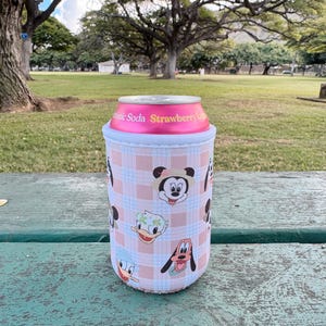 May include: A pink and white plaid can cooler featuring cartoon character faces, including Mickey Mouse, Donald Duck, and Goofy. The cooler holds a pink soda can with the text "Strawberry L". The background shows a park setting with trees and grass.