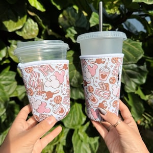 以下が含まれることがあります： 蓋と金属ストローが付いた2つの透明なプラスチックカップ。それぞれに模様入りのスリーブが付いています。スリーブは、ピンクのミッキーマウスの頭、ポップコーン、その他のテーマグラフィックを備えた水色の背景です。