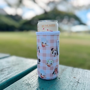 May include: A pink and white plaid can cooler featuring cartoon character faces. The cooler holds a can with the text "BE THE ALOHA" printed on it. The cooler is on a green picnic table.