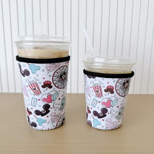 以下が含まれることがあります： 白い背景に、ミッキーマウスの頭、風船、ポップコーン、アイスクリーム、観覧車など、カラフルなパターンが描かれた、断熱された飲料用スリーブ2つ。スリーブはネオプレン製で、上部に黒いバンドが付いています。