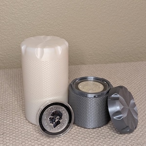 May include: A tall, off-white cylindrical container with a textured diamond pattern and a star-shaped top, alongside a shorter, dark gray cylindrical container with a matching texture, a removable lid, and a silver coin featuring a portrait.