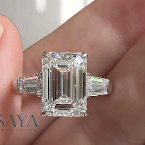 Puede incluir: Un anillo de platino con un gran diamante central de talla esmeralda, flanqueado por dos diamantes baguette más pequeños. El anillo está sostenido por una mano. La marca "SAYA ORNAMENTS" es visible.
