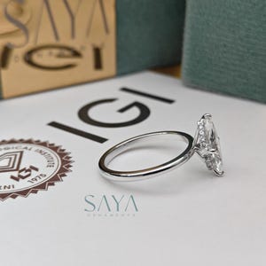 Puede incluir: Un anillo de diamantes con corte marquesa y banda plateada descansa sobre una superficie blanca. El anillo presenta un diamante grande y transparente. El logotipo de IGI y el texto "SAYA ORNAMENTS" son visibles.
