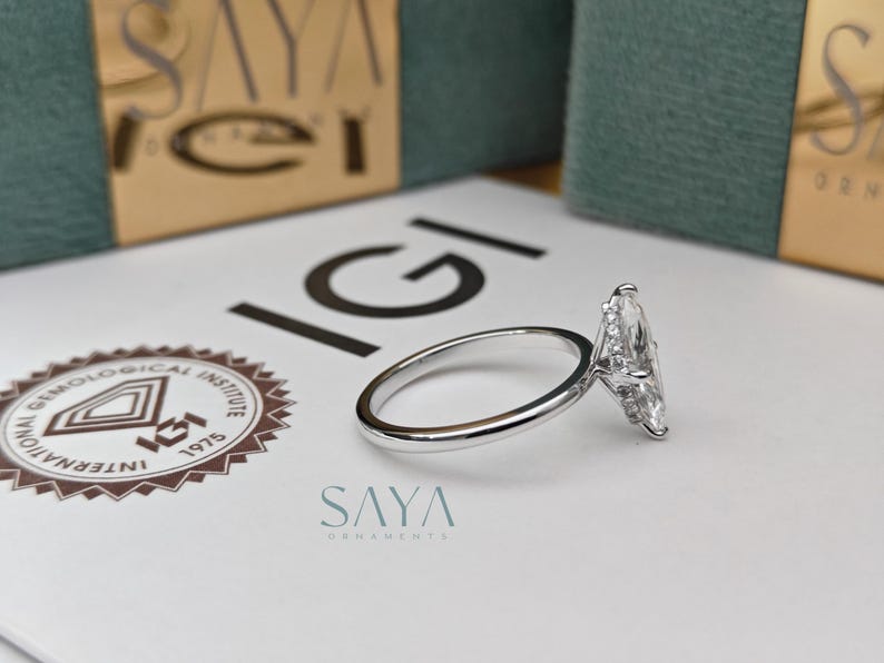 Puede incluir: Un anillo de diamantes con corte marquesa y banda plateada descansa sobre una superficie blanca. El anillo presenta un diamante grande y transparente. El logotipo de IGI y el texto "SAYA ORNAMENTS" son visibles.