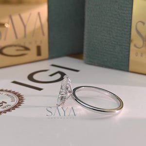 Puede incluir: Primer plano de un anillo de compromiso de diamantes con una piedra central en forma de pera. El anillo est&aacute; sobre una superficie blanca con el texto "SAYA ORNAMENTS" y un logotipo. La banda del anillo es plateada.