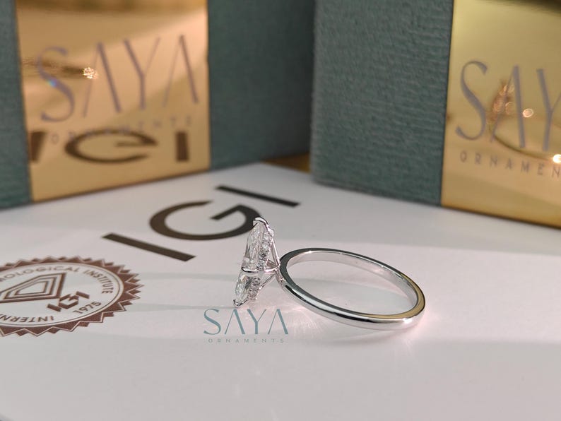 Puede incluir: Primer plano de un anillo de compromiso de diamantes con una piedra central en forma de pera. El anillo est&aacute; sobre una superficie blanca con el texto "SAYA ORNAMENTS" y un logotipo. La banda del anillo es plateada.