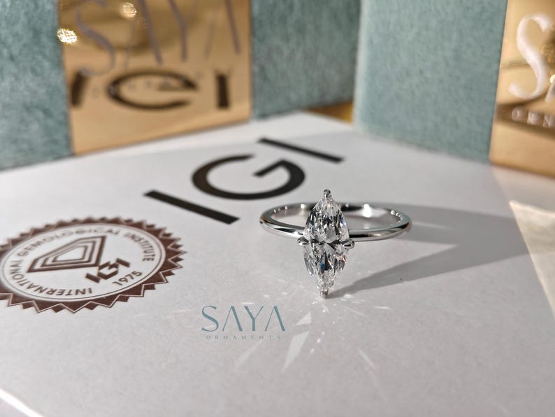 May include: A marquise-cut diamond ring with a slim silver band. The ring is on a white surface with the text "IGI" and the logo of the International Gemological Institute. The brand name "SAYA ORNAMENTS" is also visible.