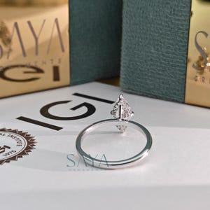 Puede incluir: Un anillo de plata con una parte superior en forma de pera con diamantes. El anillo est&aacute; sobre una superficie blanca con el texto "IGI" y "SAYA ORNAMENTS" visibles. Dos cajas doradas con el texto "SAYA ORNAMENTS" est&aacute;n en el fondo.