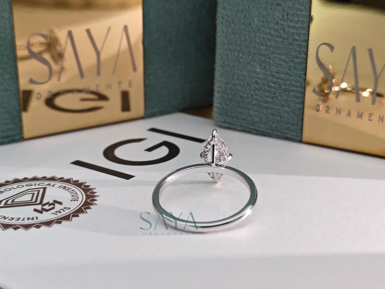 Puede incluir: Un anillo de plata con una parte superior en forma de pera con diamantes. El anillo est&aacute; sobre una superficie blanca con el texto "IGI" y "SAYA ORNAMENTS" visibles. Dos cajas doradas con el texto "SAYA ORNAMENTS" est&aacute;n en el fondo.
