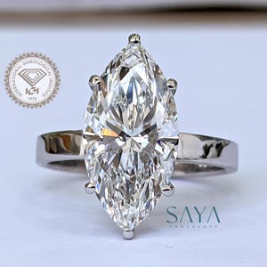 May include: A marquise-cut diamond ring with a polished silver band. The large, clear diamond is the focal point, reflecting light. The ring is set on a white surface. The logo "SAYA ORNAMENTS" is visible.
