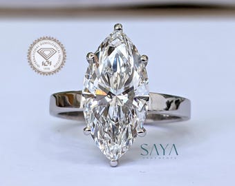 Könnte beinhalten: Ein Ring mit einem Diamanten im Marquiseschliff und einem polierten Silberband. Der große, klare Diamant ist der Mittelpunkt und reflektiert das Licht. Der Ring liegt auf einer weißen Oberfläche. Das Logo "SAYA ORNAMENTS" ist sichtbar.