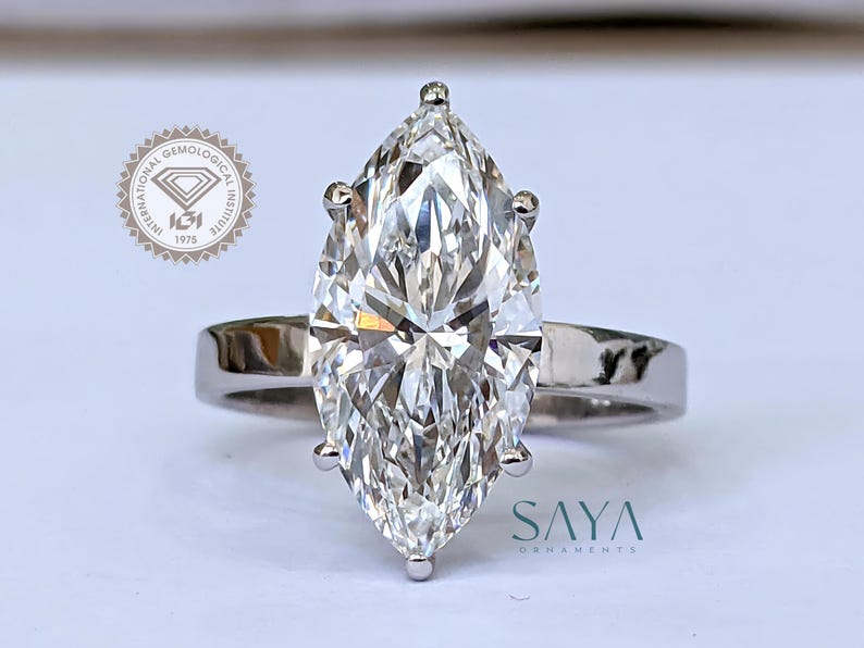 May include: A marquise-cut diamond ring with a polished silver band. The large, clear diamond is the focal point, reflecting light. The ring is set on a white surface. The logo "SAYA ORNAMENTS" is visible.
