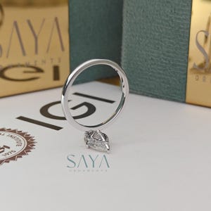 Puede incluir: Un anillo de plata con un colgante de diamante en forma de pera. El anillo se muestra sobre una superficie blanca con el logotipo "SAYA ORNAMENTS" y una caja dorada en el fondo.