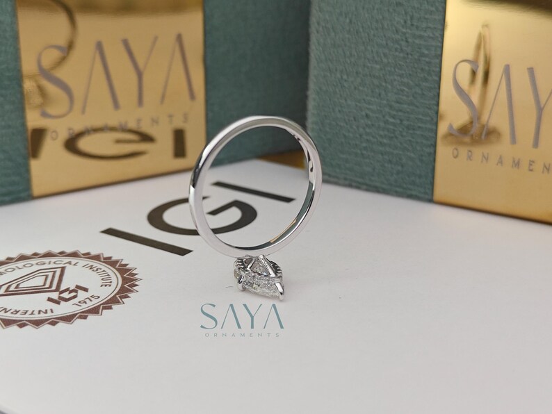 Puede incluir: Un anillo de plata con un colgante de diamante en forma de pera. El anillo se muestra sobre una superficie blanca con el logotipo "SAYA ORNAMENTS" y una caja dorada en el fondo.